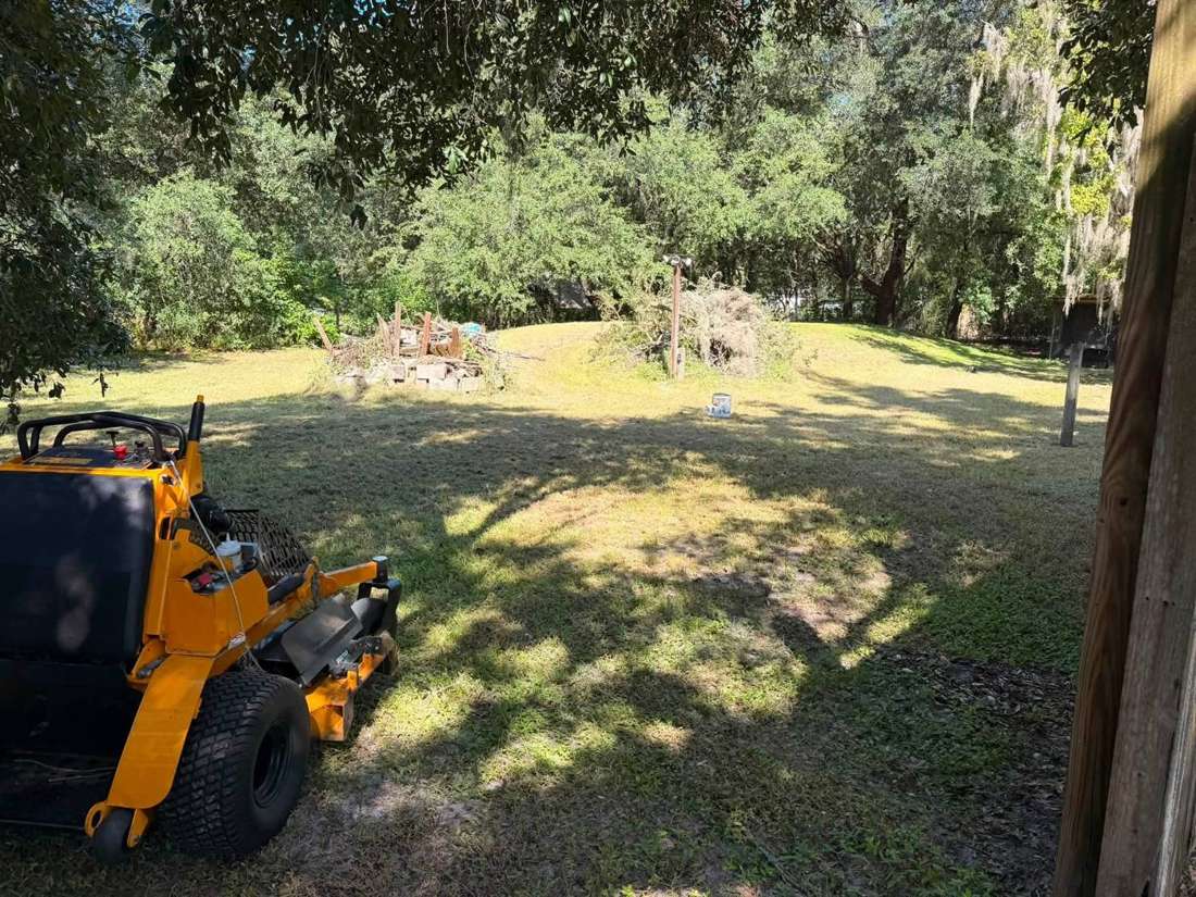 Yellow stand-on mower parked on a freshly cut shaded back yard with brush piles gathered for removal